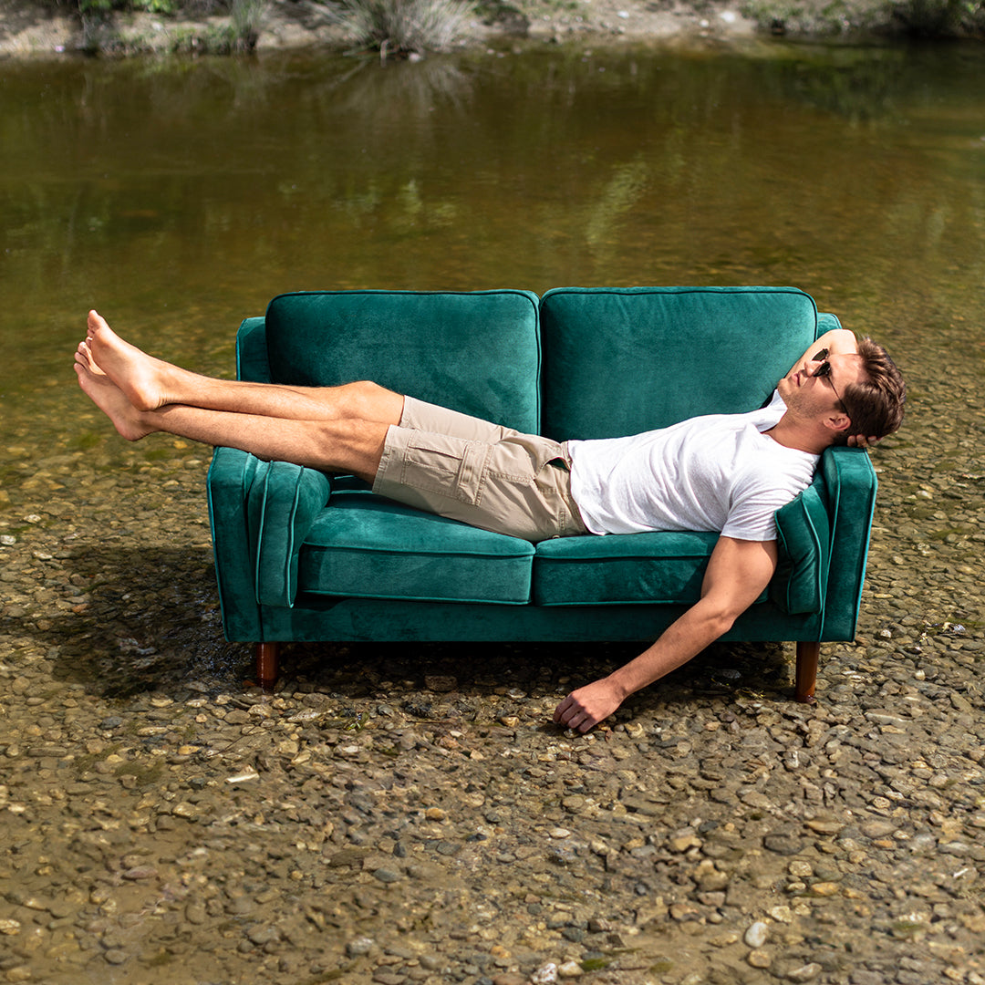 Ein junger Mann liegt entspannt auf einem grünen Sofa, das mitten im Wasser an einem Flussufer auf einem kiesigen Boden steht. Der Mann trägt ein weißes T-Shirt und beige Shorts, hat die Augen geschlossen und wirkt relaxed.