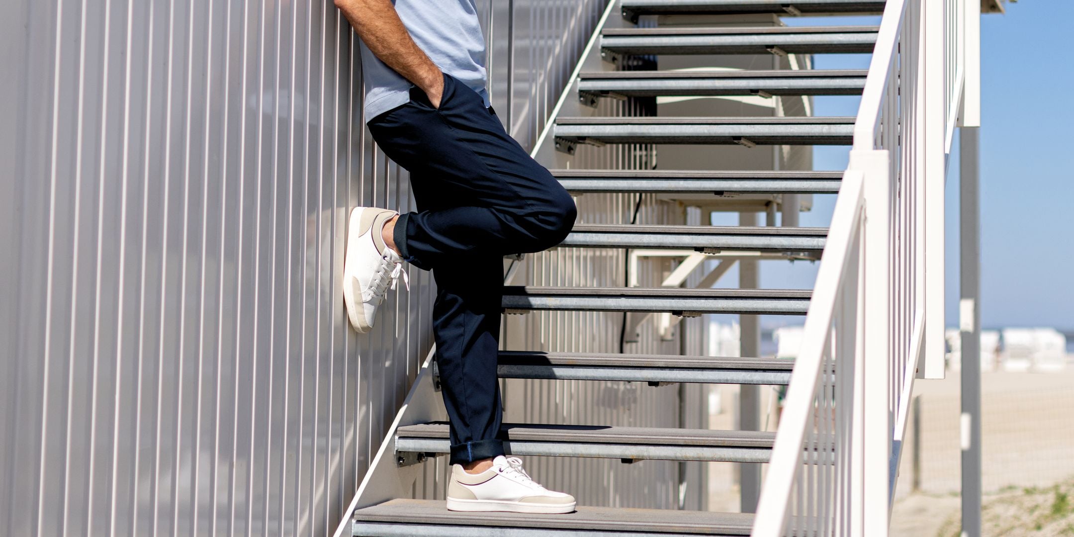 Nahaufnahme eines Mannes, der auf einer Metalltreppe im Freien steht. Er trägt dunkelblaue Hosen, weiße Sneakers und ein dunkelblaues Oberteil. Seine Arme sind zusätzlich sichtbar, während er die Treppe hinauf oder hinunter steigt. Im Hintergrund ist ein blauer Himmel und eine Strandkulisse sichtbar.