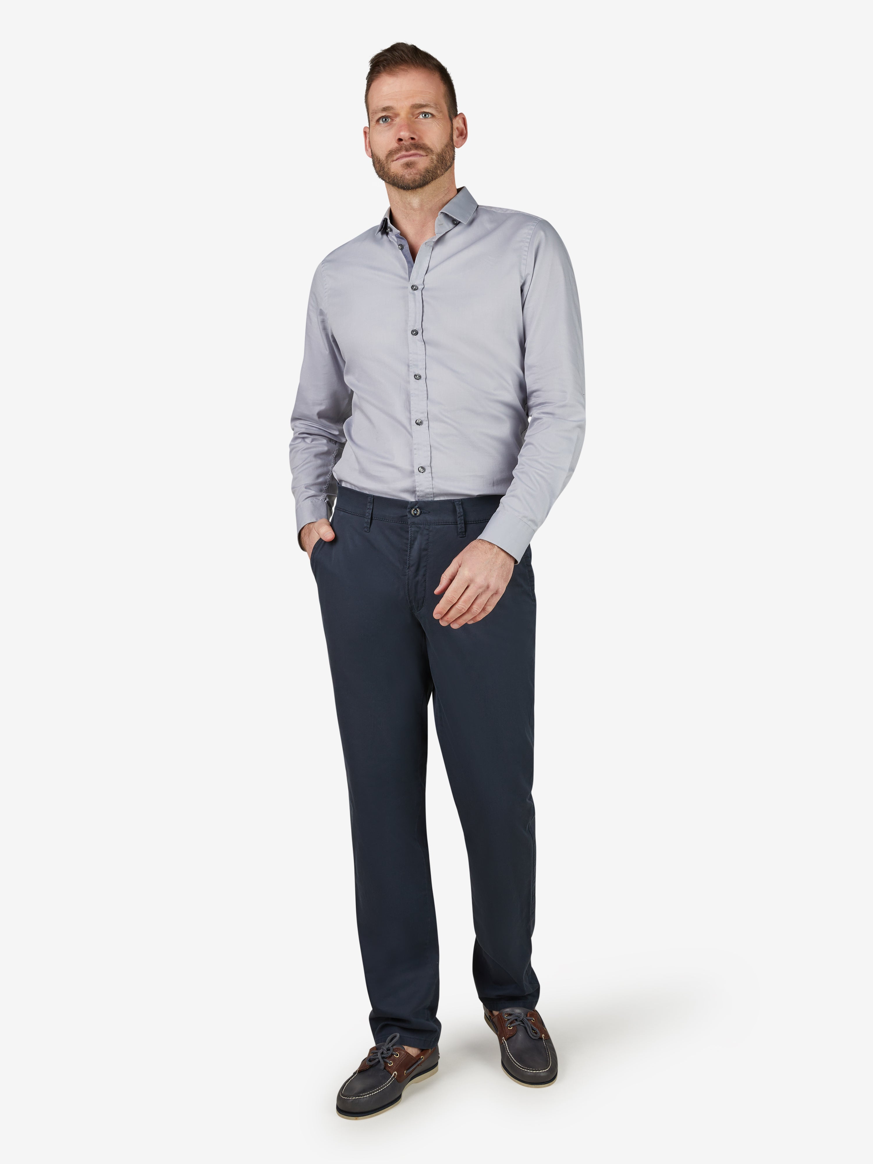 Mann in einem hellblauen Hemd und dunklen Slim-Fit Baumwoll-Chinohose mit Komfortbund, Hemdenstopper und High Stretch Qualität, in einem weißen Studio-Setup.