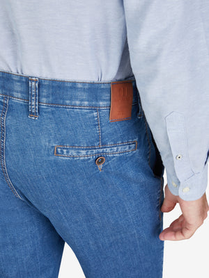 Nahaufnahme einer jeansblauen Flat Front-Hose mit elastischem Komfortbund und Hemdenstopper, gefertigt aus hoch elastischem Super High Stretch-Denim mit Lycra dual-FX®-Technologie, normaler Leibhöhe, schmaler Beinverlauf, getragen von einer Person in hellblitem Hemd, Hersteller Club of Comfort.