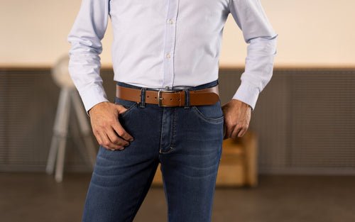 Ein Mann steht mit legerer Haltung in einem Raum, trägt eine hellblaue, langärmlige Hemd und dunkelblaue Jeans. Seine Hände sind in die Hosentaschen gesteckt, und er trägt einen braunen Ledergürtel. Im Hintergrund sind Möbel und ein Sitzbereich sichtbar.
