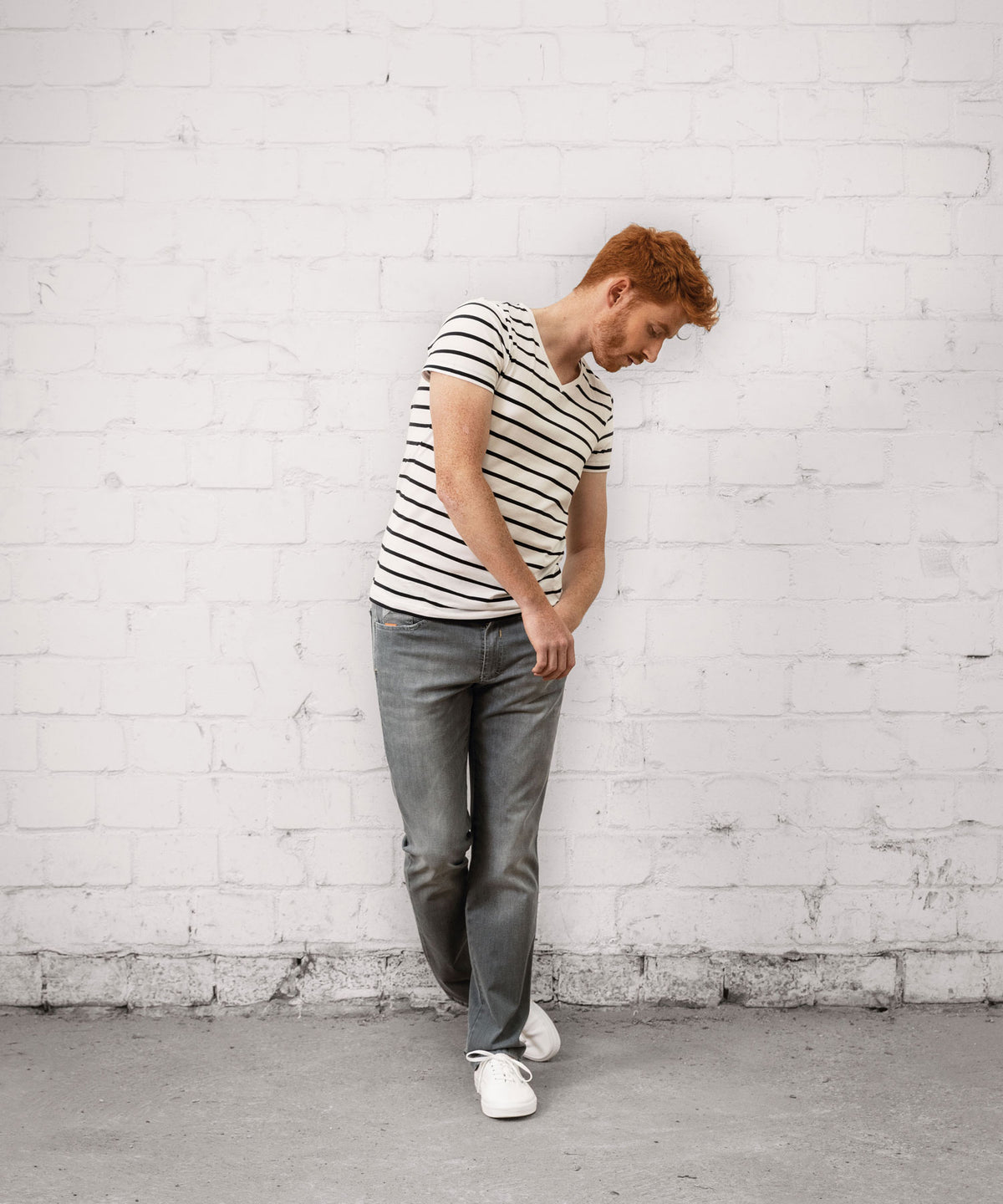 Ein junger Mann mit roten Haaren steht vor einer weißen Ziegelwand, trägt ein schwarz-weiß gestreiftes T-Shirt, graue Jeans und weiße Sneakers. Er schaut nach unten und hält seinen rechten Oberschenkel mit beiden Händen, möglicherweise zur Unterstützung oder bei Schmerzen.
