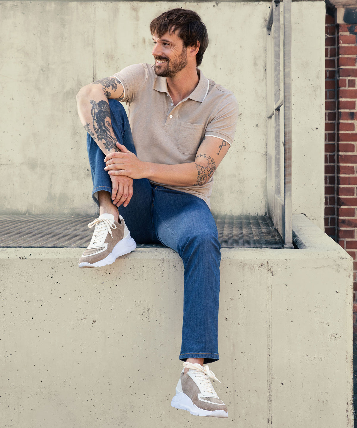 Ein lächelnder Mann mit dunklem, mittellangem Haar sitzt entspannt an einer Betonstufe vor einer urbanen Wand. Er trägt ein beige Polo-Shirt, blaue Jeans und beige Turnschuhe. Seine Arme sind tätowiert, und er schaut nach rechts.
