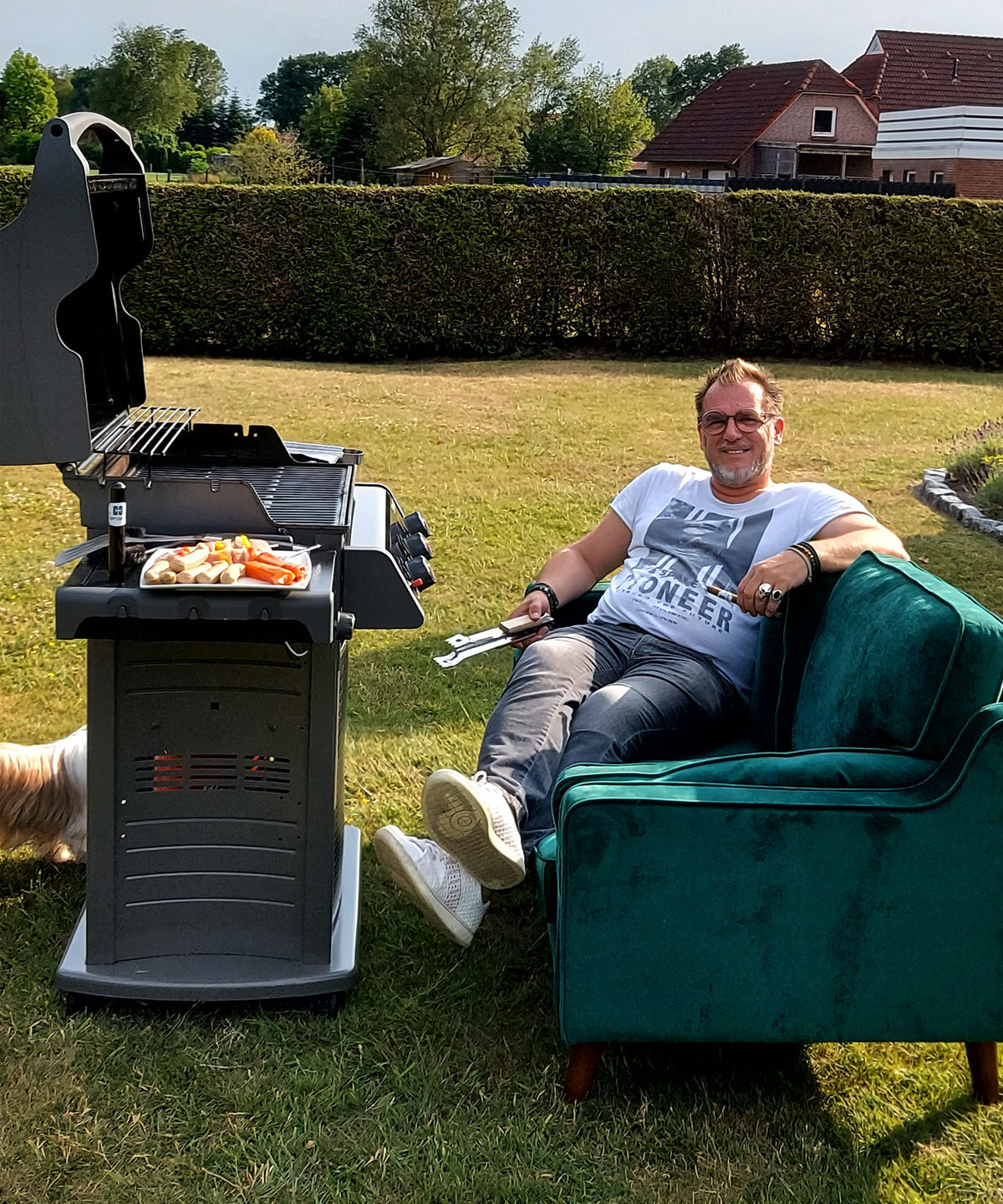 Ein lachender Mann sitzt entspannt auf einem grünen Sofa im Garten, neben einem Gasgrill mit gegrillten Würstchen und Pommes. Er hält Grillzangen in der Hand und trägt ein weißes T-Shirt mit Aufdruck. Im Hintergrund sind Bäume, eine Hecke und einige Häuser zu sehen.
