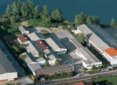 Luftaufnahme eines Gewerbegebiets mit mehreren Gebäuden, darunter Lagerhallen und Werkstätten, umgeben von Bäumen und einem Wasserarm im Hintergrund. Ein Straßenbereich mit parkenden Autos verläuft im Vordergrund.