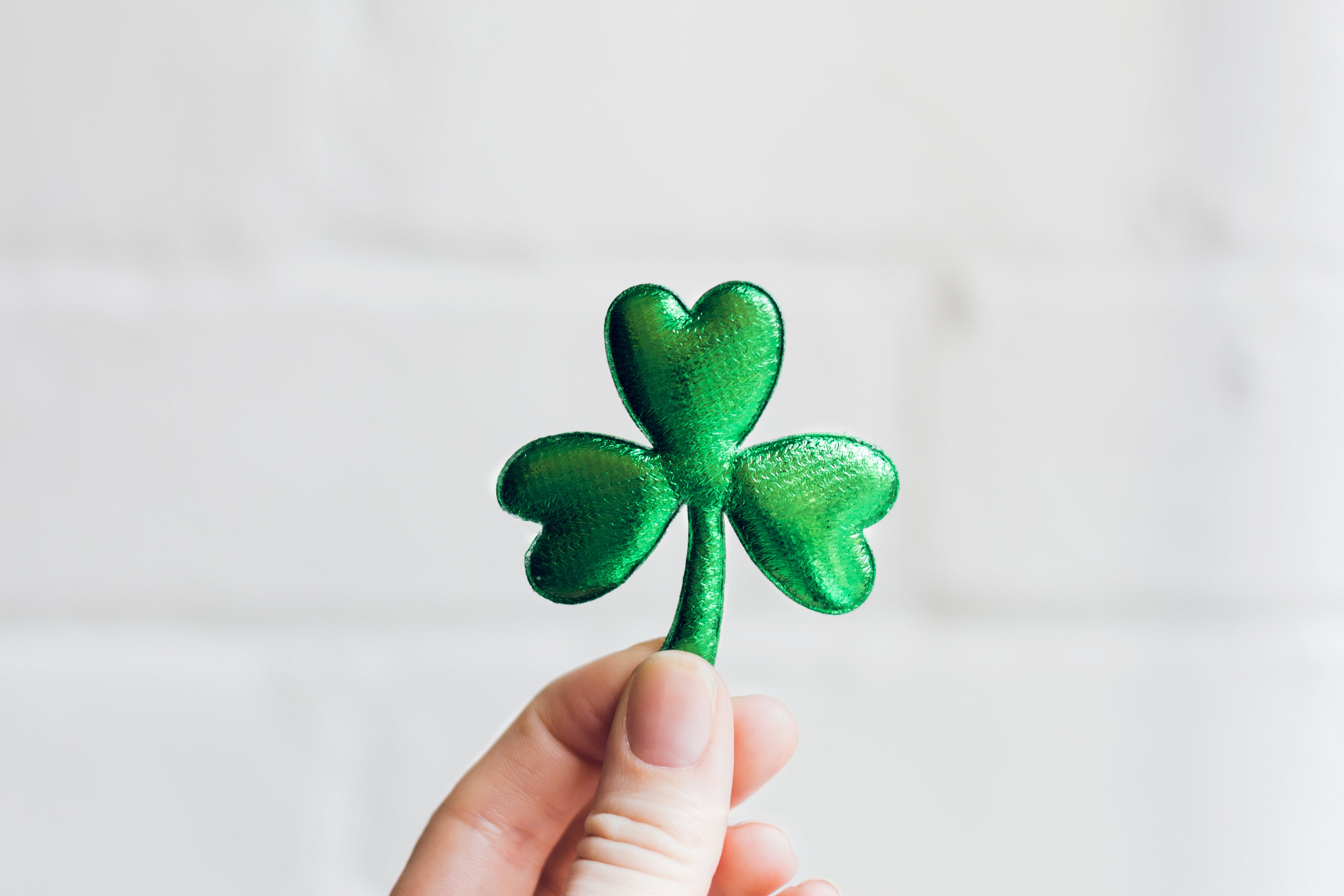 Eine Hand hält einen grünen, glitzernden Shamrock aus Filz oder Stoff vor einem weißen Hintergrund.